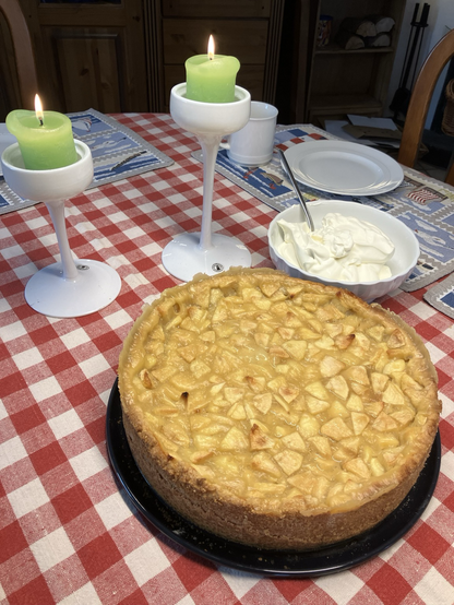 Apfel-Wein-Torte, eine Schüssel Sahne und zwei grüne Kerzen auf weißen Leuchtern, auf rot-weiß-kariertem Tischtuch